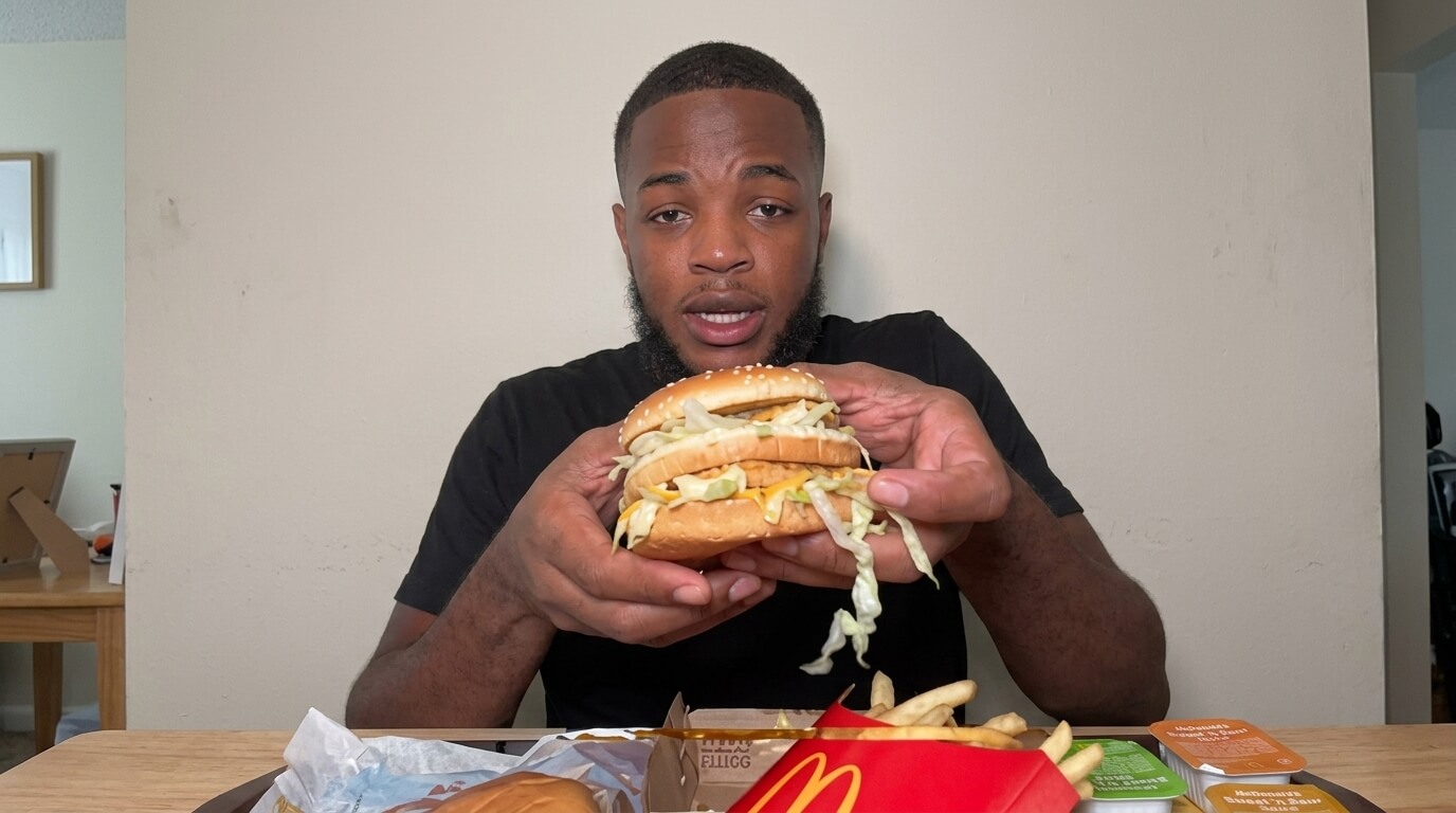 Man holding a McDonald's Big Mac burger with fries and sauces on the table in front of him