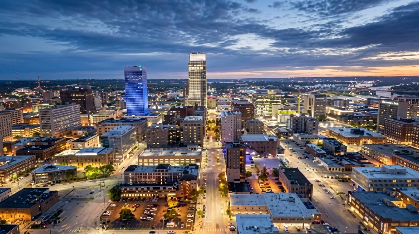 Downtown Tulsa skyline at dusk with illuminated buildings and streets under a cloudy sky.