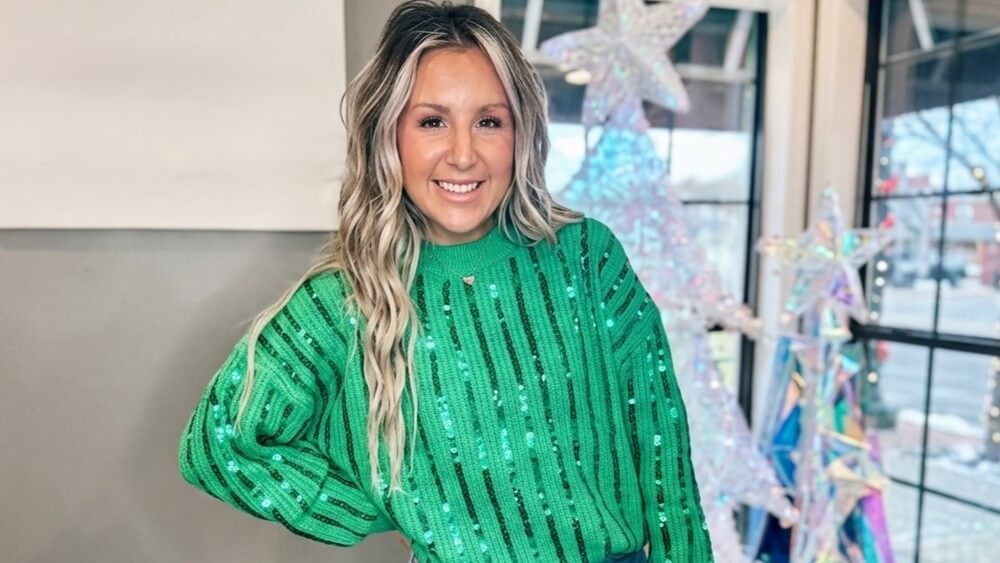 Smiling woman with long blonde hair wearing a green sequined sweater standing indoors near iridescent star decorations.
