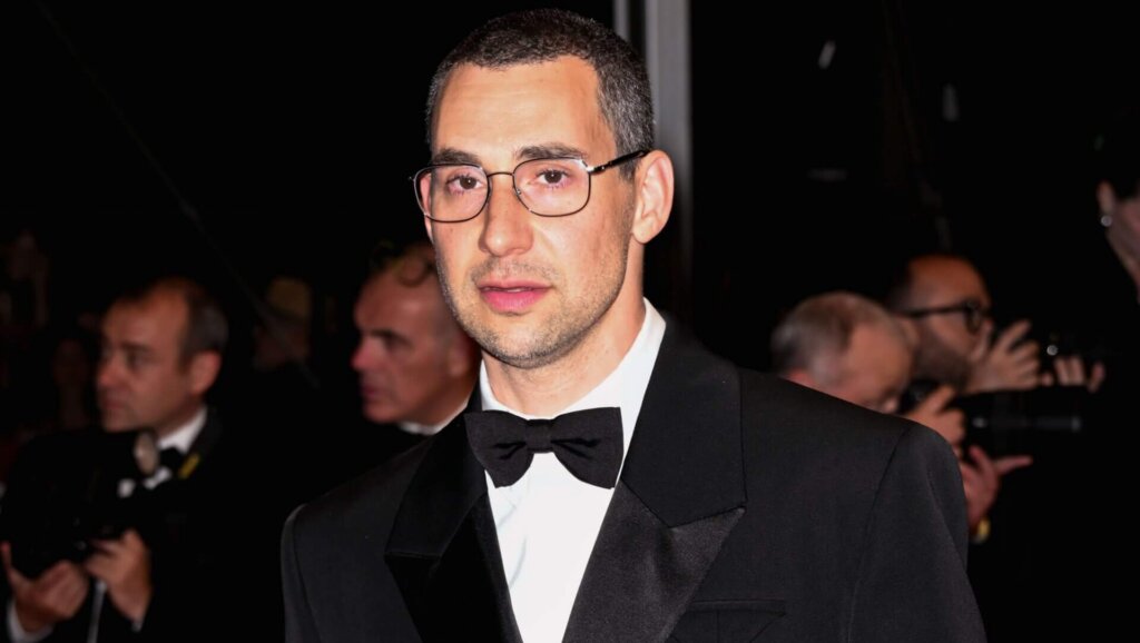 Simon Rex wearing a black tuxedo and glasses on a red carpet with photographers in the background