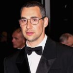 Simon Rex wearing a black tuxedo and glasses on a red carpet with photographers in the background