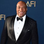 John Singleton smiling in a black tuxedo at an AFI event with a blue backdrop.