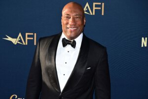 John Singleton smiling in a black tuxedo at an AFI event with a blue backdrop.