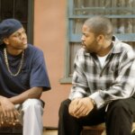 Ice Cube and Chris Tucker sitting and talking outside a house, Ice Cube in plaid shirt, Chris Tucker in navy shirt and cap