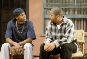 Ice Cube and Chris Tucker sitting and talking outside a house, Ice Cube in plaid shirt, Chris Tucker in navy shirt and cap