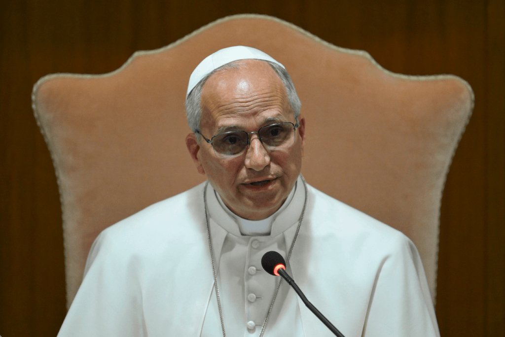 Pope Francis speaking into a microphone while seated on a beige chair against a wooden background