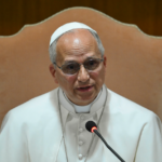 Pope Francis speaking into a microphone while seated on a beige chair against a wooden background