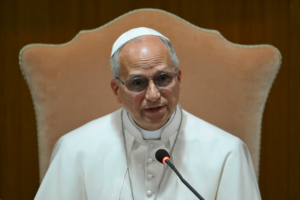 Pope Francis speaking into a microphone while seated on a beige chair against a wooden background