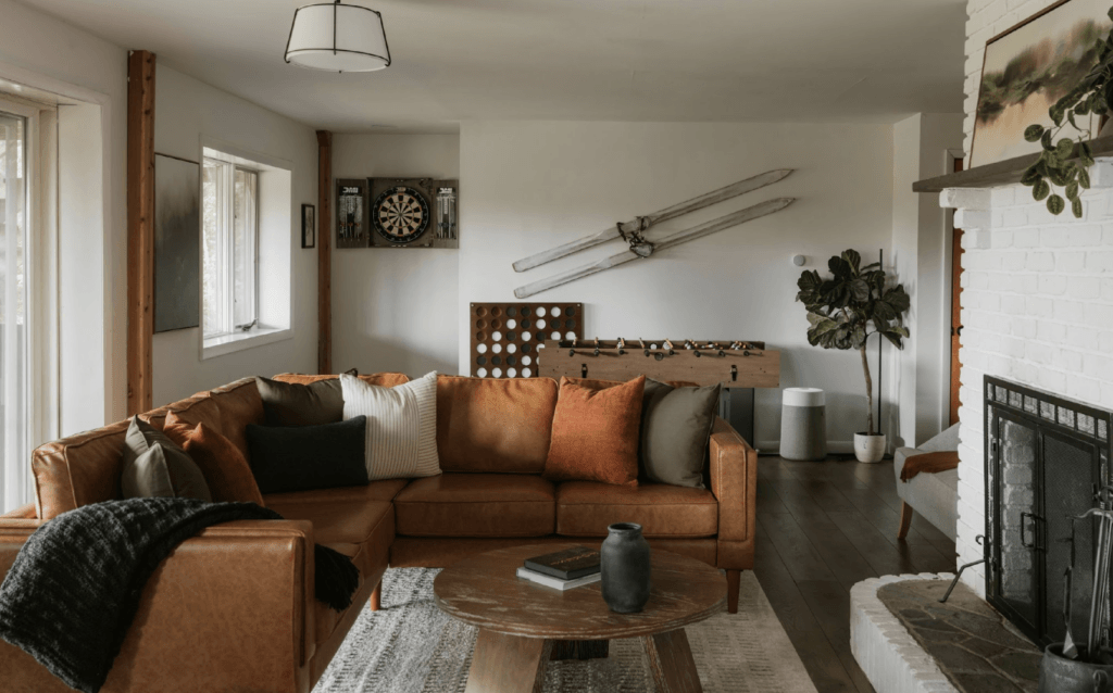 Living room with brown leather sectional sofa, dartboard, foosball table, vintage skis on wall, and white brick fireplace