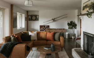 Living room with brown leather sectional sofa, dartboard, foosball table, vintage skis on wall, and white brick fireplace