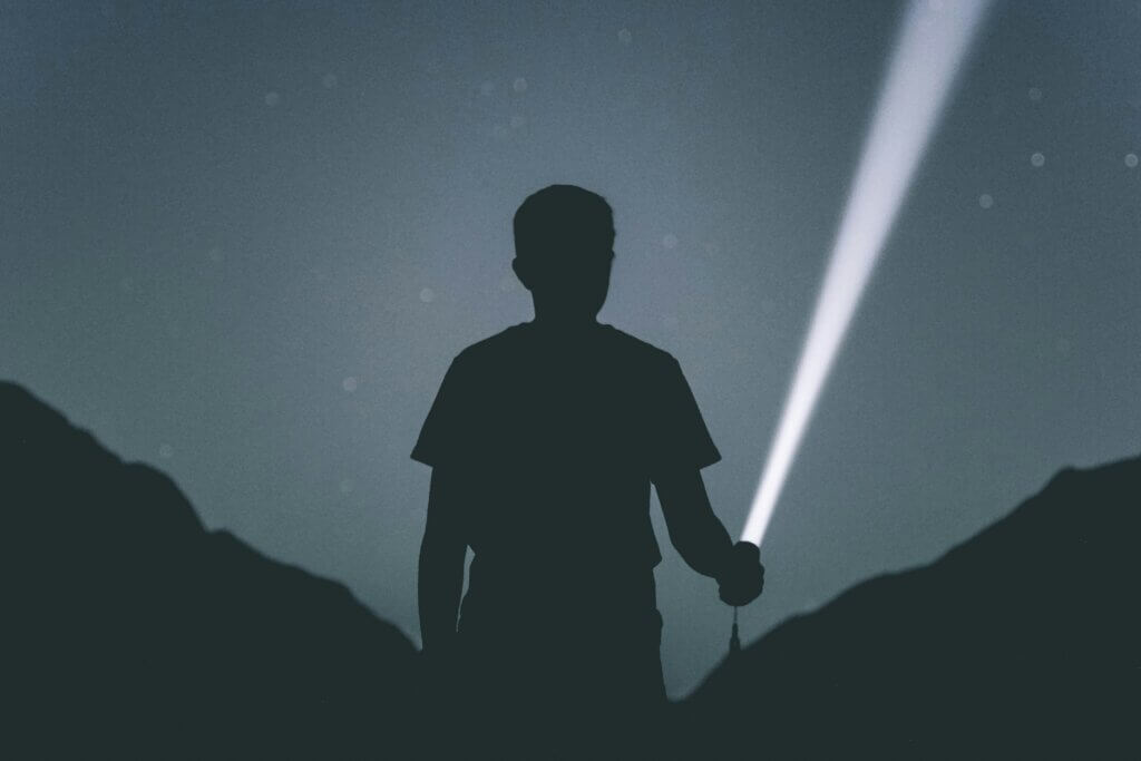Silhouette of a person holding a flashlight shining a beam of light in a dark outdoor setting at night.