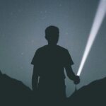 Silhouette of a person holding a flashlight shining a beam of light in a dark outdoor setting at night.