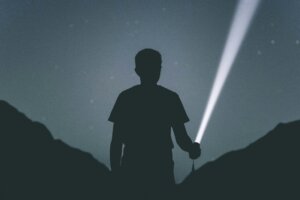 Silhouette of a person holding a flashlight shining a beam of light in a dark outdoor setting at night.