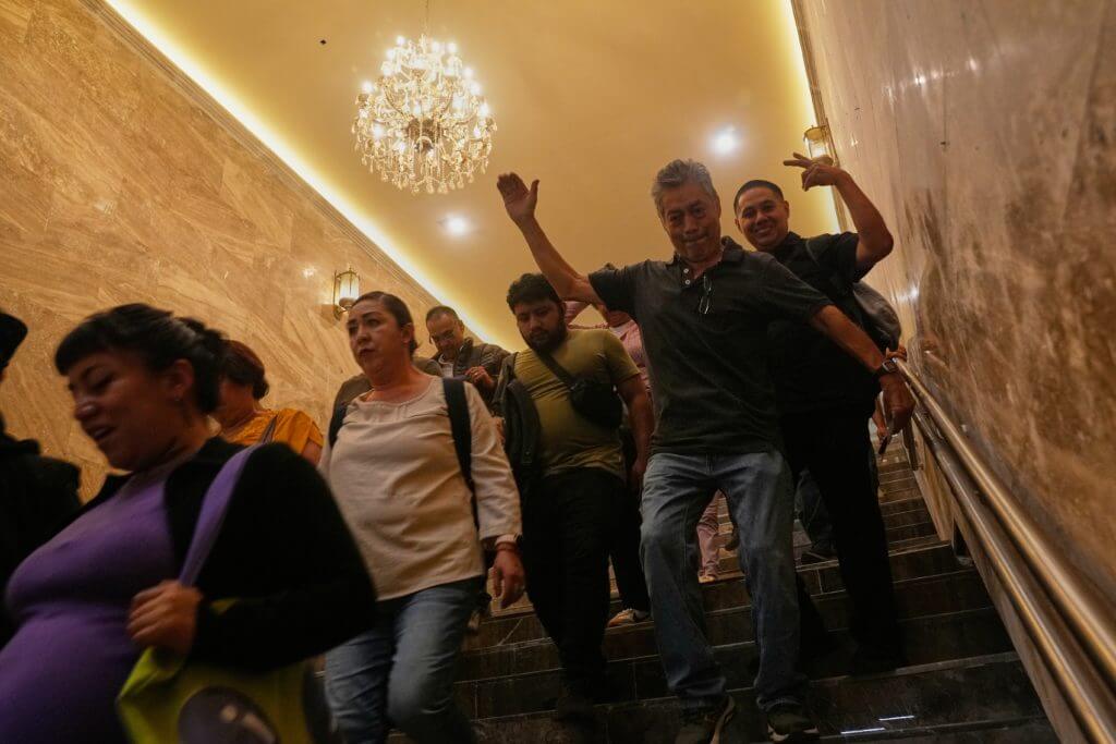 Group of people walking down a marble staircase under a chandelier, with one man raising his hand and smiling