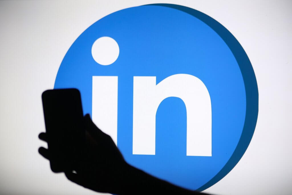 Silhouette of a hand holding a smartphone in front of the LinkedIn logo on a screen