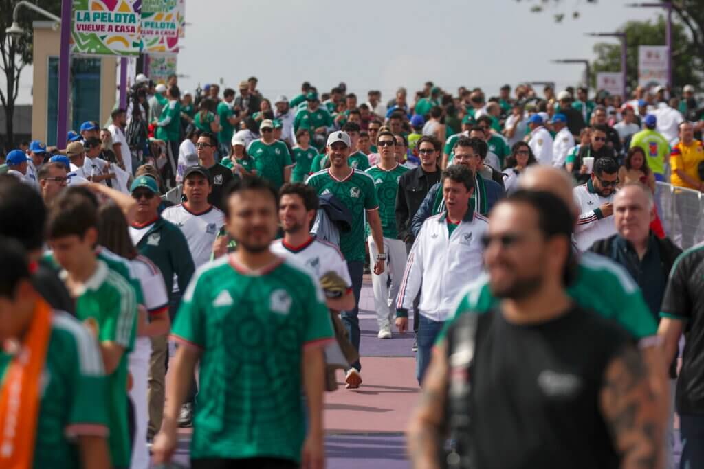 Crowd of soccer fans wearing Mexico national team jerseys walking along a street with banners reading "La Pelota Vuelve a Casa"