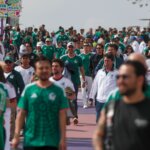 Crowd of soccer fans wearing Mexico national team jerseys walking along a street with banners reading "La Pelota Vuelve a Casa"