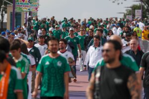 Crowd of soccer fans wearing Mexico national team jerseys walking along a street with banners reading "La Pelota Vuelve a Casa"