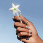 Hand with silver nail art and rings holding a white star-shaped wand with a green gem against a blue sky background
