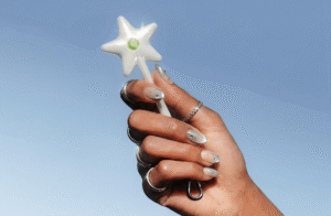 Hand with silver nail art and rings holding a white star-shaped wand with a green gem against a blue sky background
