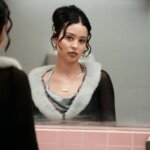 Young woman with dark hair in a bun wearing a black top with white fur trim looking at herself in a bathroom mirror