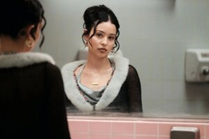 Young woman with dark hair in a bun wearing a black top with white fur trim looking at herself in a bathroom mirror