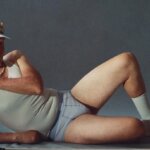 Man in white tank top, shorts, and visor lying on side with golf clubs and ball on gray background