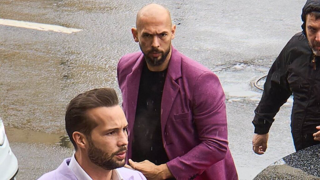 Man in lavender suit adjusting collar with another man in purple blazer and black shirt behind him on wet street