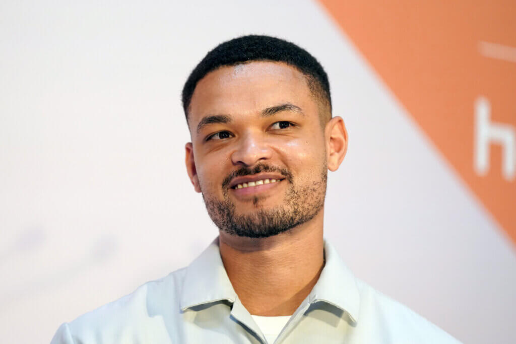 Close-up of a smiling man with short hair and a trimmed beard wearing a light-colored collared shirt against a white and orange background.