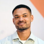 Close-up of a smiling man with short hair and a trimmed beard wearing a light-colored collared shirt against a white and orange background.