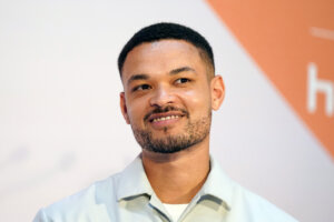 Close-up of a smiling man with short hair and a trimmed beard wearing a light-colored collared shirt against a white and orange background.