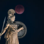 Statue of Athena holding a spear and shield with a blood moon in the night sky behind it