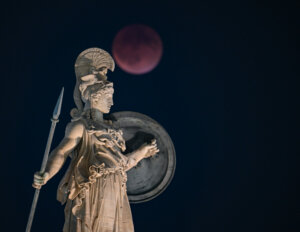Statue of Athena holding a spear and shield with a blood moon in the night sky behind it