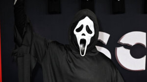 Person wearing Ghostface costume from Scream holding a knife with multiple Ghostface masks in the background