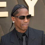 Travis Scott wearing sunglasses, diamond earrings, and a pinstripe suit at an event with gold letters in the background