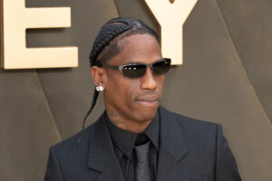 Travis Scott wearing sunglasses, diamond earrings, and a pinstripe suit at an event with gold letters in the background