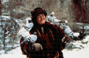Woman in winter clothing smiling with pigeons perched on her arms and shoulders in a snowy outdoor setting