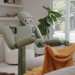 Home robot folding an orange towel in a living room with large windows and plants.