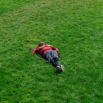 Person lying on green grass wearing a red and black striped sweater and dark pants.