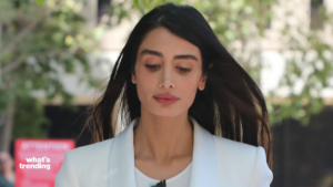 Woman with long dark hair wearing a white blazer outdoors with blurred greenery in the background