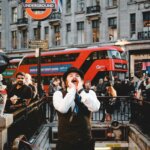 Man in vintage outfit shouting near London Underground entrance with red double-decker bus and crowd in background