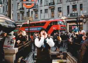 Man in vintage outfit shouting near London Underground entrance with red double-decker bus and crowd in background