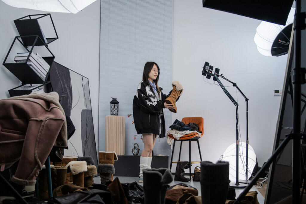 Woman holding a fur-lined brown boot in a studio surrounded by multiple boots and professional lighting equipment.