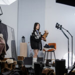 Woman holding a fur-lined brown boot in a studio surrounded by multiple boots and professional lighting equipment.