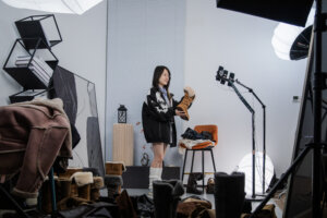 Woman holding a fur-lined brown boot in a studio surrounded by multiple boots and professional lighting equipment.