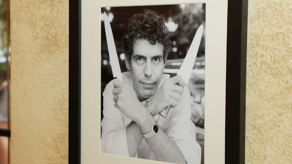Black and white framed photo of a man holding two knives crossed in front of his face on a textured wall.