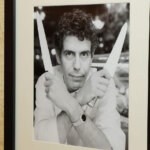 Black and white framed photo of a man holding two knives crossed in front of his face on a textured wall.