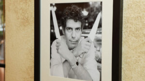 Black and white framed photo of a man holding two knives crossed in front of his face on a textured wall.
