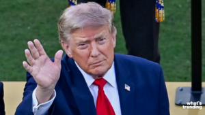 Donald Trump in a blue suit and red tie waving with a serious expression outdoors