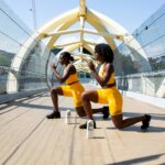Two women in yellow workout outfits doing lunges on a pedestrian bridge with Boxed Water cartons nearby